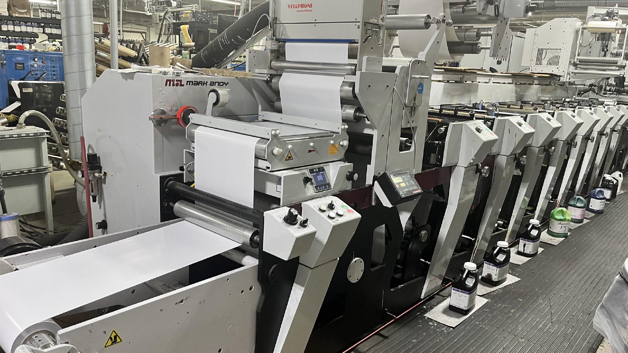 A Mark Andy P7 flexo press line with ink stations running the length of the deck and a corona treatment unit mounted above the web